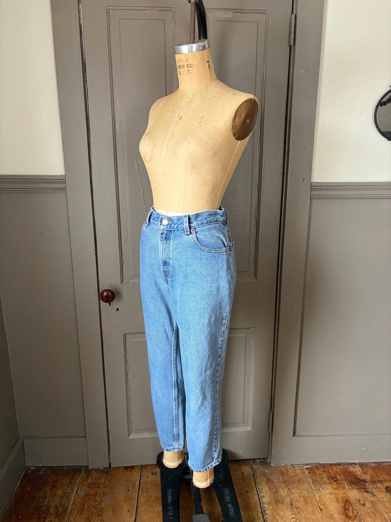 May include: Light blue denim jeans displayed on a vintage dress form. The jeans have a classic five-pocket design, a button and zipper closure, and a straight leg fit. The jeans are a medium wash and appear to be in good condition.