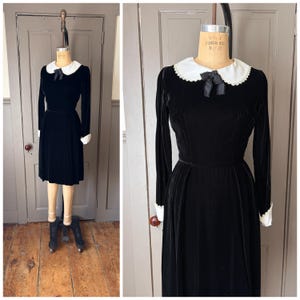 Puede incluir: Un vestido de terciopelo negro con cuello y puños blancos de estilo Peter Pan. El vestido de manga larga presenta un corpiño entallado y una falda a la rodilla. Un lazo negro adorna el cuello. El vestido se exhibe en un maniquí vintage.