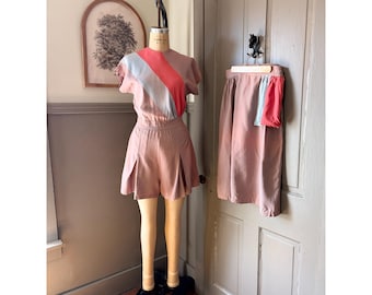 May include: A two-piece set consisting of a short-sleeved top and pleated shorts in a dusty rose color. The top features a diagonal color-blocked design with light blue and coral stripes. A matching skirt with a light blue and coral accent panel is included.