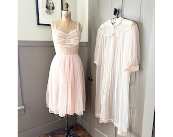 Puede incluir: Un conjunto de lencería rosa pálido de estilo vintage y una bata a juego. La lencería presenta un delicado ribete de encaje y una falda de tul. La bata transparente tiene cuello y cierre de botones, colgada de una puerta.
