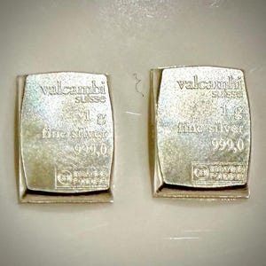 May include: Two rectangular silver bars, each weighing 1g. The text on each bar reads: valcambi suisse, 1 g, fine silver, 999,0, along with a small logo at the bottom. The bars have a metallic, reflective surface and are positioned side by side.