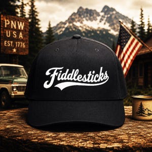 May include: A black baseball cap featuring the word Fiddlesticks embroidered in white script lettering across the front panel. The cap has a structured crown, a curved brim, and a button at the top center, showcasing a clean and simple design.