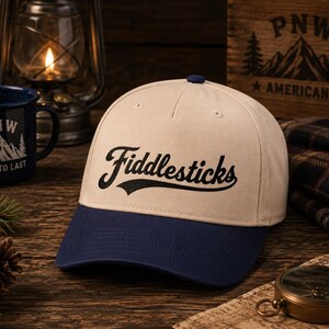 May include: A two-tone baseball cap featuring a cream-colored crown and a dark blue brim. The front of the cap displays the word Fiddlesticks embroidered in a bold, black, cursive script font. The cap has a structured five-panel design.