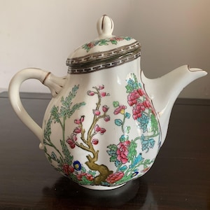 Op de afbeelding: Een witte keramische theepot met een handgeschilderd bloemmotief in roze, groene en blauwe tinten. De theepot heeft een gebogen handvat, een tuit en een bijpassend deksel met een geometrisch bruin randpatroon op zowel de deksel als de rand.