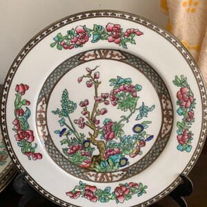 Könnte beinhalten: Ein runder weißer Keramikteller mit einer zentralen Abbildung eines blühenden Baumes mit rosa und blauen Blüten. Der Rand ist mit einem geometrischen Muster sowie floralen Motiven in Rosa, Grün und Blau verziert und mit Gold akzentuiert.