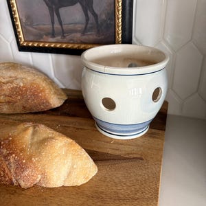 May include: A cream-coloured ceramic garlic keeper with a rounded body, featuring multiple circular ventilation holes and a blue horizontal band near the base. The surface has a visible crackle glaze finish and a wide opening at the top.