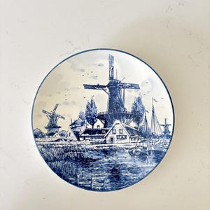 May include: A round ceramic plate featuring a blue and white transferware design of a Dutch landscape. The scene depicts two windmills, a small house, and two sailboats on a canal, all rendered in detailed blue ink on a cream-coloured background.