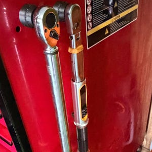 Puede incluir: Dos llaves dinamométricas de metal con pantallas digitales y empuñaduras de goma amarilla están fijadas al lateral de un armario de herramientas rojo. Las llaves tienen ejes de cromo plateado, cabezales negros y botones de control.