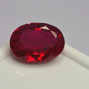 May include: A vibrant, deep red gemstone with an oval shape and a faceted cut. The stone displays multiple geometric facets that reflect light, creating a brilliant appearance with varying shades of crimson and dark red throughout the surface.