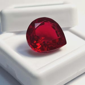 Könnte beinhalten: Ein birnenförmiger, facettierter Edelstein in einer tiefen, leuchtend roten Farbe. Der Stein liegt auf der Oberfläche einer weißen, rechteckigen Kunststoff-Aufbewahrungsbox mit Klappdeckel und zeigt seine klare, reflektierende Facettenstruktur.