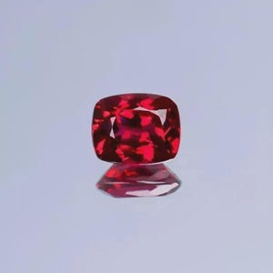 Puede incluir: Una piedra preciosa de talla cojín con un color rojo intenso y vibrante y una superficie facetada que refleja la luz. La piedra muestra una apariencia clara y pulida con bordes definidos y una forma simétrica, proyectando un reflejo.