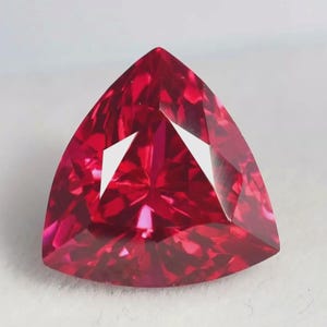 Può includere: Una gemma rosso vivo e profondo tagliata in forma triangolare con molteplici sfaccettature. La pietra presenta una superficie brillante e riflettente con bordi netti e una qualità trasparente che cattura la luce in modo nitido.