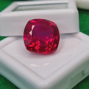 Può includere: Una gemma rosso intenso e vibrante con taglio a cuscino e superficie sfaccettata, appoggiata su una scatola portaoggetti in plastica bianca. La pietra mostra un'intensa saturazione e chiarezza, riflettendo la luce sulle sue facce geometriche.