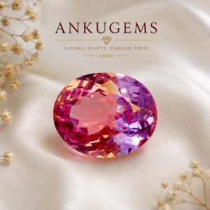 Könnte beinhalten: Ein oval geschliffener Edelstein mit einem lebendigen Farbverlauf in Pink, Violett und Orange. Der Text ANKUGEMS, ein Diamantsymbol und der Schriftzug NATURAL BEAUTY, TIMELESS TRUST sind über dem auf weißem Stoff liegenden Stein abgebildet.