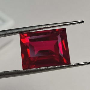 May include: A vibrant, deep red gemstone with a rectangular emerald cut, featuring sharp, clean facets that reflect light. The stone is held securely between the two thin, metallic tips of a pair of silver-toned tweezers, showcasing its clarity.
