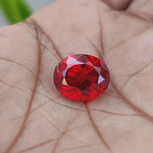 May include: A vibrant, oval-cut gemstone with a deep, saturated red color. The stone features a faceted surface that reflects light, creating a brilliant, sparkling appearance. The gemstone is displayed in a clear, polished, and transparent state.