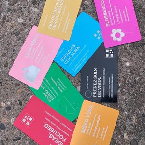 Op de afbeelding: Een verzameling van zeven rechthoekige visitekaartjes in verschillende kleuren, waaronder geel, roze, blauw, zwart, groen, rood en oranje. Elke kaart bevat unieke merknamen, contactgegevens en grafische iconen in witte of donkere tekst.