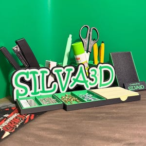 Op de afbeelding: Een groene en zwarte bureau-organizer met de tekst SILVA3D in dikke groene letters met een witte omlijning. De organizer bevat meerdere vakken voor kantoorbenodigdheden zoals een nietmachine, schaar, lijm, pennen en kleine metalen onderdelen.