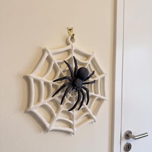 Op de afbeelding: Een witte, ronde, driedimensionale spinnenweb wanddecoratie met een opvallende, massief zwarte spin in het midden. Het web heeft een dik, gegoten frame met een klein lusje aan de bovenkant om het aan een haak aan de muur op te hangen.