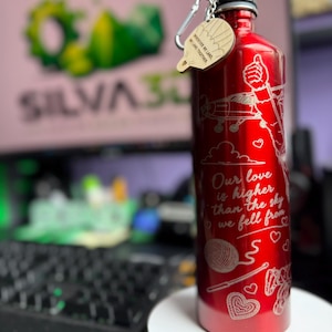 Op de afbeelding: Een metallic rode waterfles met witte lasergegraveerde illustraties van een vliegtuig, wolken, hartjes en een bol wol. De tekst luidt: "Our love is higher than the sky we fell from". Een houten label aan de dop vermeldt: "Wherever we land, we land together".