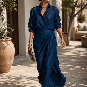 May include: A deep blue long-sleeved button-down shirt with a subtle vertical texture paired with a matching floor-length wrap skirt featuring a high side slit. The ensemble is completed with dark sunglasses, a layered necklace, and a small clutch.