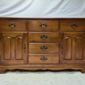 May include: A wooden buffet with a warm brown finish. It features six drawers and two cabinet doors, each with ornate brass-colored hardware. The buffet has a classic design with a slightly curved base.