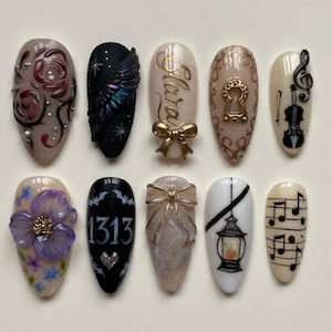 May include: A set of ten press-on nails featuring diverse designs including 3D roses, a black wing, the name Clara with a gold bow, a gold keyhole, a violin, a purple flower, the number 1313 with bats, a textured bow, a lantern, and musical notes.