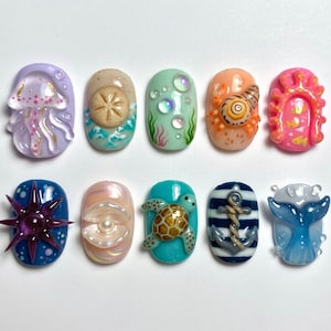 May include: A set of ten oval-shaped 3D nail art charms featuring ocean-themed designs including a jellyfish, sand dollar, bubbles, crab, coral, starfish, pearl in a shell, sea turtle, anchor, and whale tail in various colors and textures.