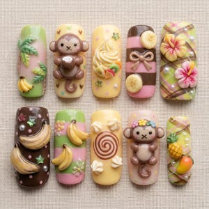 May include: A set of ten decorative nail art tips featuring 3D designs including brown monkeys, yellow bananas, tropical hibiscus flowers, pineapples, mangoes, whipped cream swirls, and brown stripes on pastel green, yellow, and brown backgrounds.