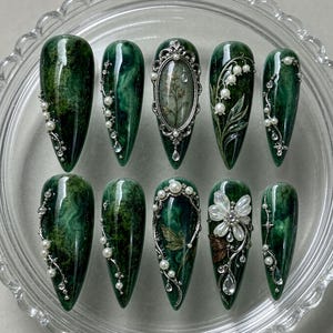 May include: A set of ten stiletto-shaped press-on nails featuring a deep forest green marbled finish. The nails are decorated with silver metal filigree frames, white pearl accents, clear rhinestones, and detailed botanical illustrations of flowers.