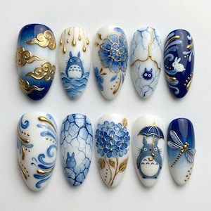 May include: A set of ten almond-shaped press-on nails featuring intricate blue and white designs with gold accents. The artwork includes depictions of a cartoon creature, floral patterns, cracked textures, a dragonfly, and swirling clouds.