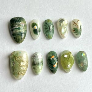 May include: A set of ten press-on nails featuring various green-toned designs, including marbled patterns, a small white flower, gold leaf accents, a painted koi fish, a pressed green leaf, and small iridescent rhinestones on a white background.