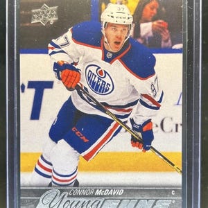 May include: A Connor McDavid Young Guns hockey trading card encased in a clear plastic sleeve. The card features an action shot of the player in a white, blue, and orange Edmonton Oilers jersey, holding a hockey stick on the ice rink.