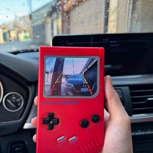 Puede incluir: Una consola de juegos portátil de color rojo brillante con una pantalla que muestra la vista de la cámara de reversa de un automóvil con líneas de guía. El dispositivo tiene un pad direccional negro, dos botones circulares negros y dos botones plateados.