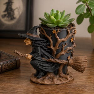 Könnte beinhalten: Ein schwarzer zylindrischer Pflanztopf mit einer skulptierten Hexe mit spitzem Hut auf einem Besenstiel. Der Boden und die Seiten sind mit braunen Baumzweigen und kleinen Blättern verziert. Eine grüne Sukkulente ist in den Topf gepflanzt.