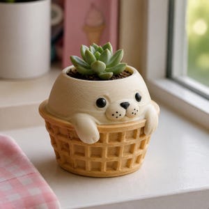 Könnte beinhalten: Ein cremefarbener Keramikübertopf in Form einer Robbe, die in einer beigen Waffelschale sitzt. Die Robbe hat schwarze Augen und eine schwarze Nase. Eine kleine grüne Sukkulente ist oben im Kopf der Robbe in dunkler Erde eingepflanzt.