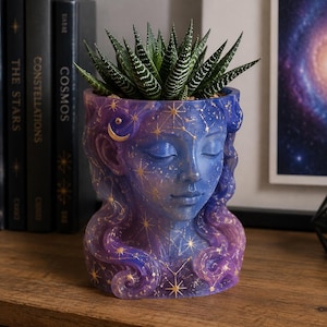 Pode incluir: Um vaso em forma de cabeça com um gradiente de tons roxos e azuis, decorado com padrões celestiais dourados, incluindo estrelas e uma lua crescente. O vaso retrata um rosto com olhos fechados e cabelo ondulado, contendo uma planta suculenta verde.
