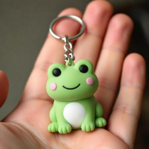 Könnte beinhalten: Ein hellgrüner Frosch-Schlüsselanhänger mit glatter, matter Oberfläche, zwei schwarzen runden Augen, einem einfachen geschwungenen schwarzen Mund, zwei kleinen rosa runden Wangen und einem weißen runden Fleck auf dem Bauch, befestigt an einem silbernen Metallring.