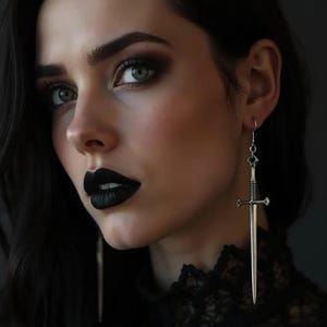 Peut inclure: Une femme aux cheveux noirs et au rouge à lèvres noir porte une boucle d'oreille argentée en forme d'épée. La boucle d'oreille est un long pendentif détaillé avec une garde croisée et une lame pointue. Le maquillage des yeux de la femme est sombre.