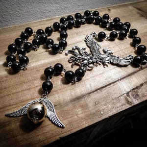 Peut inclure: Un collier de perles noires avec des breloques argentées. Les breloques comprennent un phénix et un vif d'or doré avec des ailes argentées. Le collier est présenté sur une surface en bois.