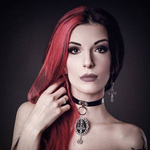 May include: A woman with red and black hair wearing a black choker necklace with a silver pendant featuring a cross design. She is also wearing silver earrings with cross charms.
