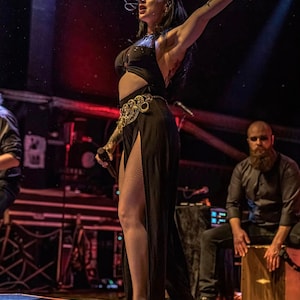 May include: A woman in a black outfit with a gold belt sings into a microphone on stage. A man with a beard sits behind a wooden percussion instrument.