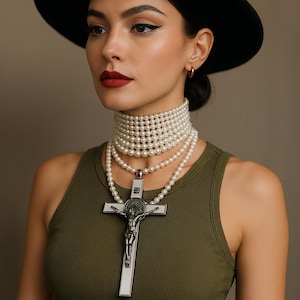 May include: A person wearing a black hat, olive green vest top, and multiple pearl necklaces. The lowest necklace has a large silver crucifix pendant. The person has dark hair and red lipstick.