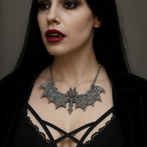 Peut inclure: Collier pendentif chauve-souris argenté avec des détails complexes. Le collier de style gothique présente un motif floral central et une chaîne. Le modèle porte un haut noir.