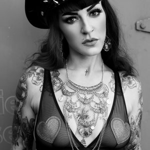 May include: A woman with black hair and a black leather cap with silver studs is wearing a black sheer top with a silver necklace and a silver cross tattoo on her neck. She has tattoos on her arms and chest, including a large eye tattoo on her stomach.