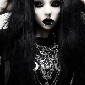 Peut inclure: Une femme aux longs cheveux noirs et au maquillage sombre porte un haut noir avec un collier argenté orné d'un pentagramme, de croissants de lune et d'autres éléments décoratifs.