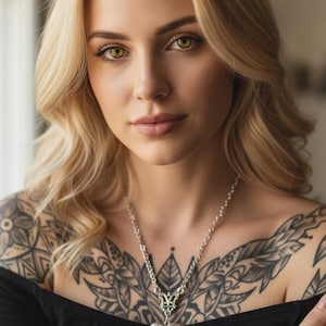 Peut inclure: Une femme aux cheveux blonds et aux yeux verts portant un haut noir à épaules dénudées. Elle a des tatouages complexes sur la poitrine et les bras, et un collier pendentif argenté en forme de cœur.