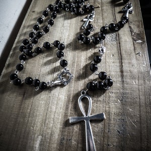 Peut inclure: Un collier de perles noires avec des pendentifs en forme d'ankh argentés. Le collier est posé sur une surface en bois.