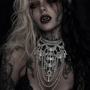 Peut inclure: Image gothique avec un collier argenté à plusieurs niveaux orné d'un pendentif ankh. La personne a des cheveux noirs et blancs, du maquillage sombre et des tatouages faciaux. L'image a une esthétique sombre et dramatique.