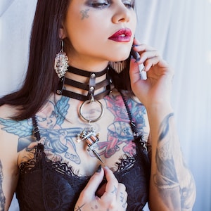 May include: A woman with multiple tattoos, wearing a black leather choker with a silver ring and a tattoo machine pendant. She is also wearing a black lace top. Her dark hair is long and straight, and she is holding a phone to her ear.